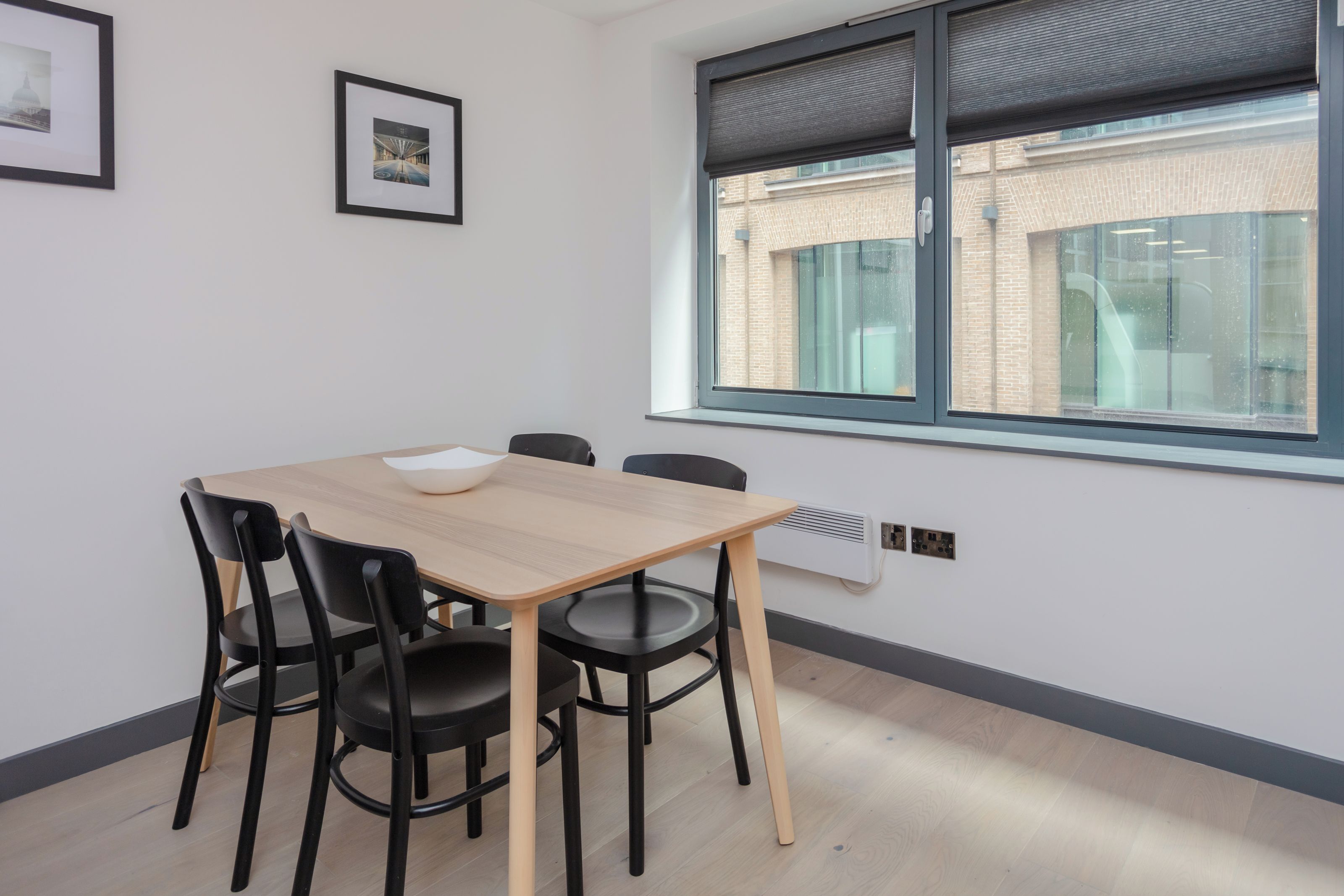 Une pièce avec une table en bois, quatre chaises noires, un bol blanc, deux tableaux encadrés accrochés au mur et une fenêtre donnant sur un bâtiment en briques.