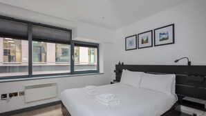 A modern hotel room with a large bed, a window with blinds, a bedside table, and a wall-mounted lamp. - Centrally Located 1BD Flat in the Heart of London (London)