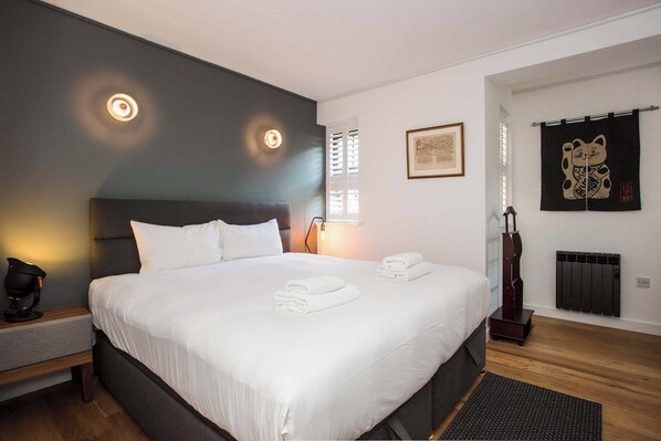 A modern hotel room with a large bed, bedside tables, a nightstand, a wall-mounted lamp, a framed picture, a decorative wall hanging, and a radiator.