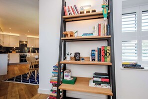 A modern kitchen with a dining area, a bookshelf with books and decorative items, and a window with blinds.