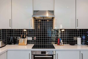 A modern kitchen with white cabinets, a black tiled backsplash, a stainless steel range hood, and a built-in oven.