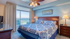 A hotel room with a large bed, a ceiling fan, a desk, a chair, a lamp, and a window with curtains. - 2BD Ocean View-Lazy River/Hot Tubs/Indoor Pool (North Myrtle Beach)