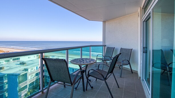 A balcony with a table and chairs, overlooking a beach and buildings. - 2BD Ocean View-Lazy River/Hot Tubs/Indoor Pool (North Myrtle Beach)