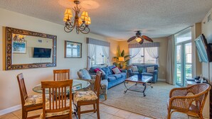 A living room with a blue sofa, wooden chairs, a glass table, a ceiling fan, and a large mirror. - 2BD Ocean View-Lazy River/Hot Tubs/Indoor Pool (North Myrtle Beach)