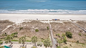 Ein Strand mit einer Holzpromenade, die zu einer sandigen Fläche mit spärlicher Vegetation und freiem Blick auf den Ozean führt.