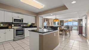 A modern kitchen with white cabinets, a central island, stainless steel appliances, and a dining area with a view. - Spectacular 3BD Corner Unit - Waterway Views - Large Square Footage (North Myrtle Beach)