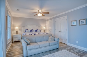 A bedroom with a bed, a ceiling fan, two bedside tables with lamps, a gray sofa, and a wooden floor.