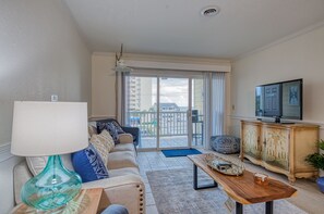 A living room with a sofa, a coffee table, a TV, and a balcony with a view of buildings. - Windy Hill-2BD Ocean View Condo - Snowbird Special (North Myrtle Beach)