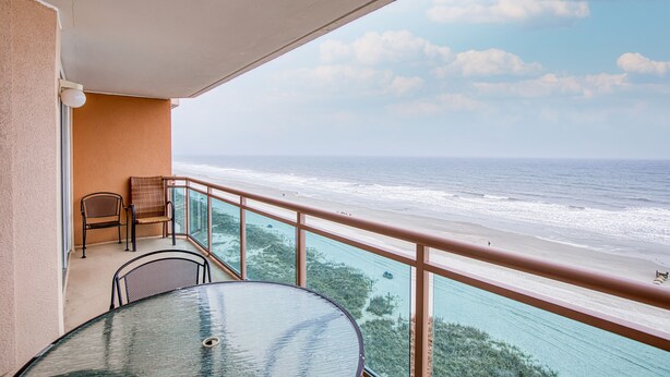 A balcony with a glass table and chairs, overlooking a beach with clear waters and a distant shoreline. - Beautiful Oceanfront-3BD-Pool/Hot Tub/Lazy River-Bahama Sands (North Myrtle Beach)