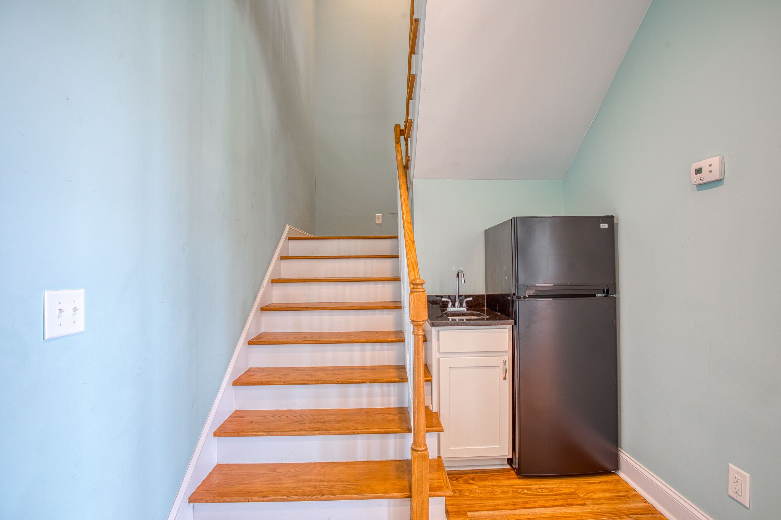 A staircase with wooden steps and a wooden handrail leading to a kitchen area with a black refrigerator and white cabinets.