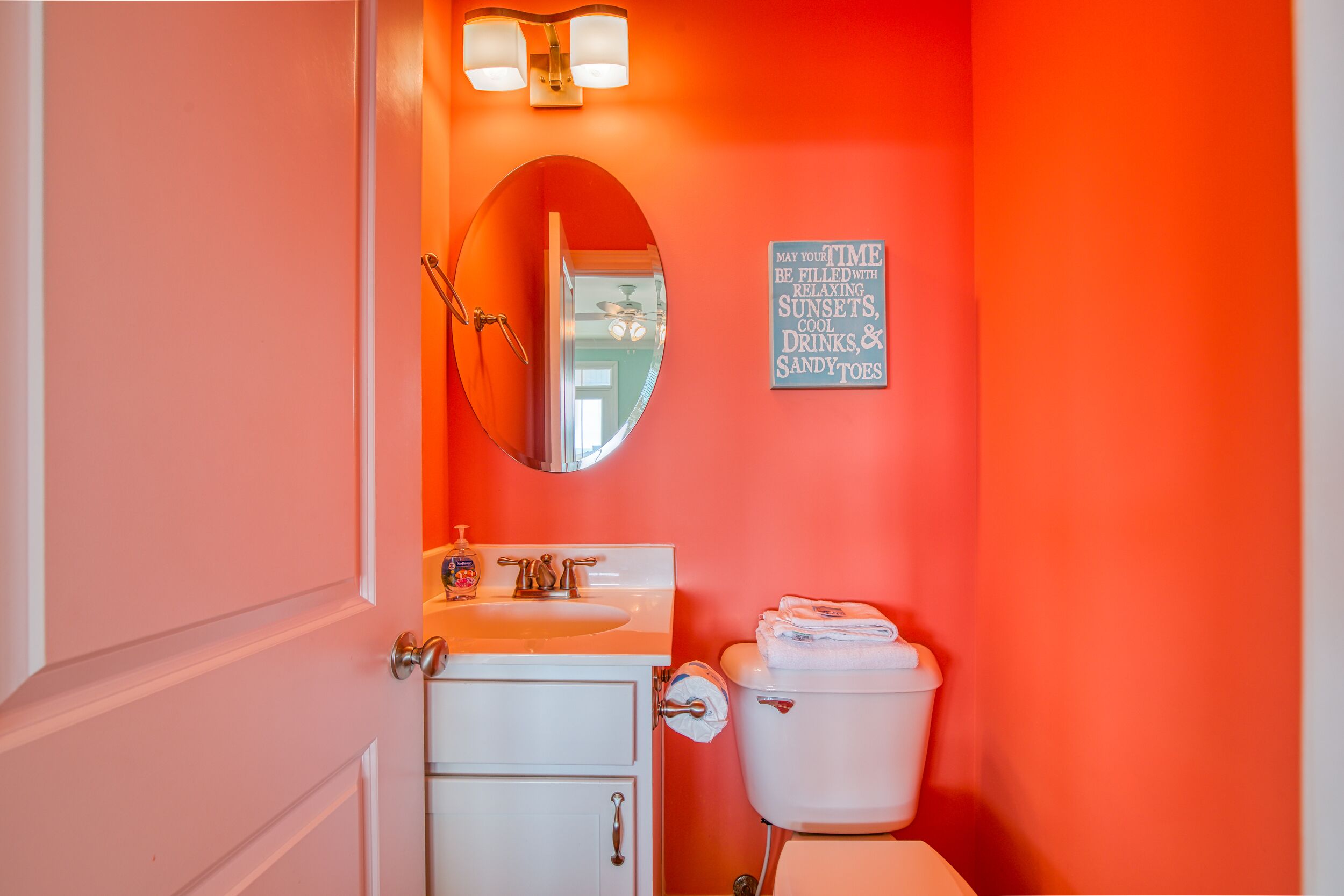 A bathroom with a white sink, a toilet, and a mirror. There is a sign on the wall that reads "May your time be filled with relaxing sunsets, cool drinks, and sandy toes."