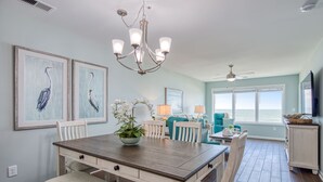 A dining area with a wooden table, white chairs, and a chandelier. There are framed artworks on the wall, a plant on the table, and a view of the ocean through large windows. - Sea Oats D-3, 3BD 2BA Oceanfront Paradise (North Myrtle Beach)