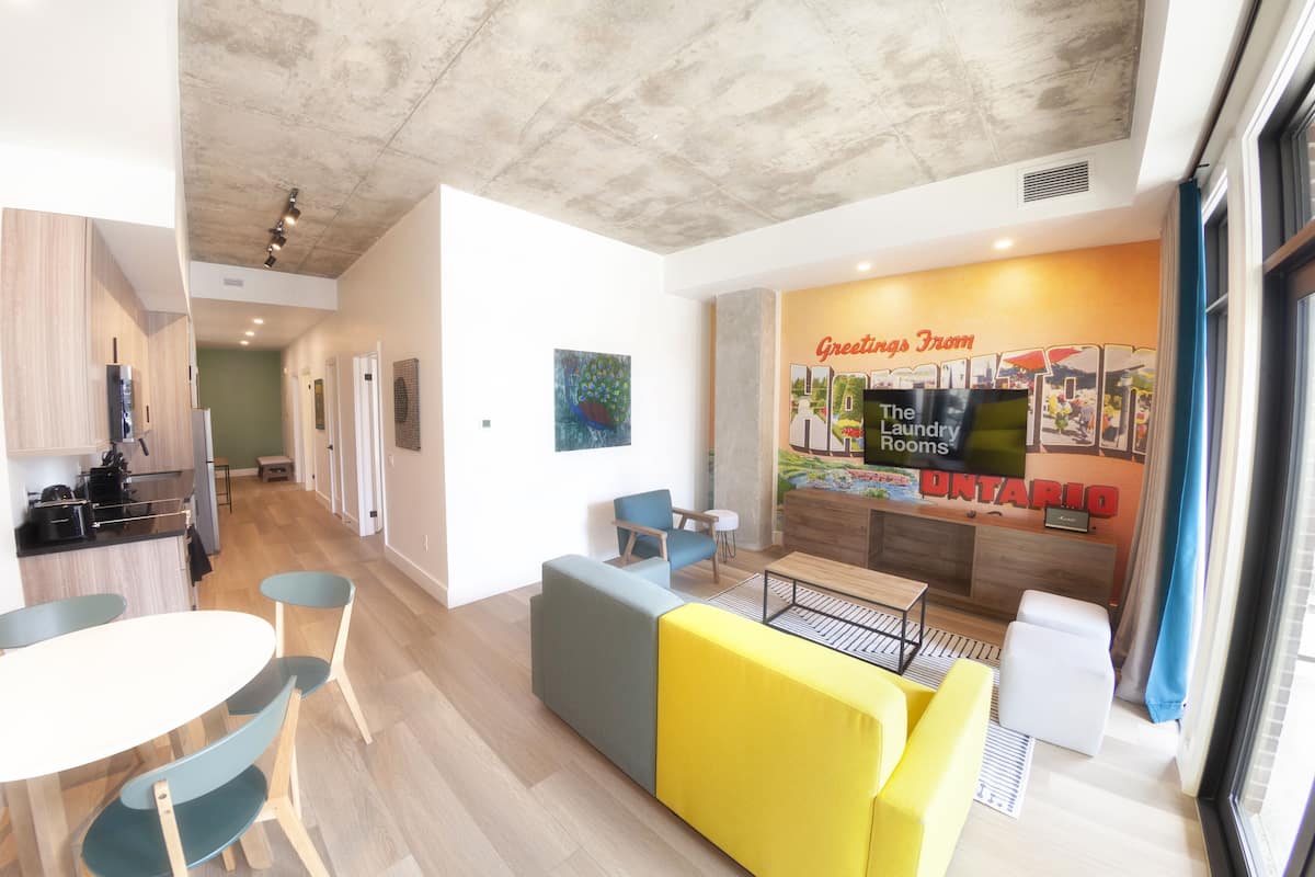 Un interior de hotel moderno con cocina, un comedor con mesa y sillas, una zona de estar con sofá y un mostrador de recepción con un cartel que dice 'Greetings from The Laundry Rooms Ontario'.