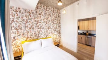 A bedroom with a bed, a wooden nightstand, a wall with a patterned wallpaper, a hanging light fixture, and a view of a kitchen with wooden cabinets and a dishwasher.