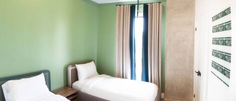 Quarto com duas camas, janela com cortinas, assoalho de madeira e porta com a placa 'HELLO'.