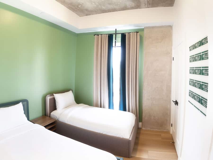 Een kamer met twee bedden, een raam met gordijnen, een houten vloer en een deur met een 'HELLO' bord.