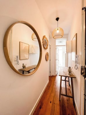 A hallway with a large mirror, wooden flooring, a small table with a vase, and a door leading to another room.