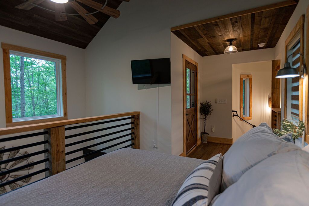A bedroom with a bed, wooden headboard, a television mounted on the wall, a window with a view of trees, and a ceiling fan.