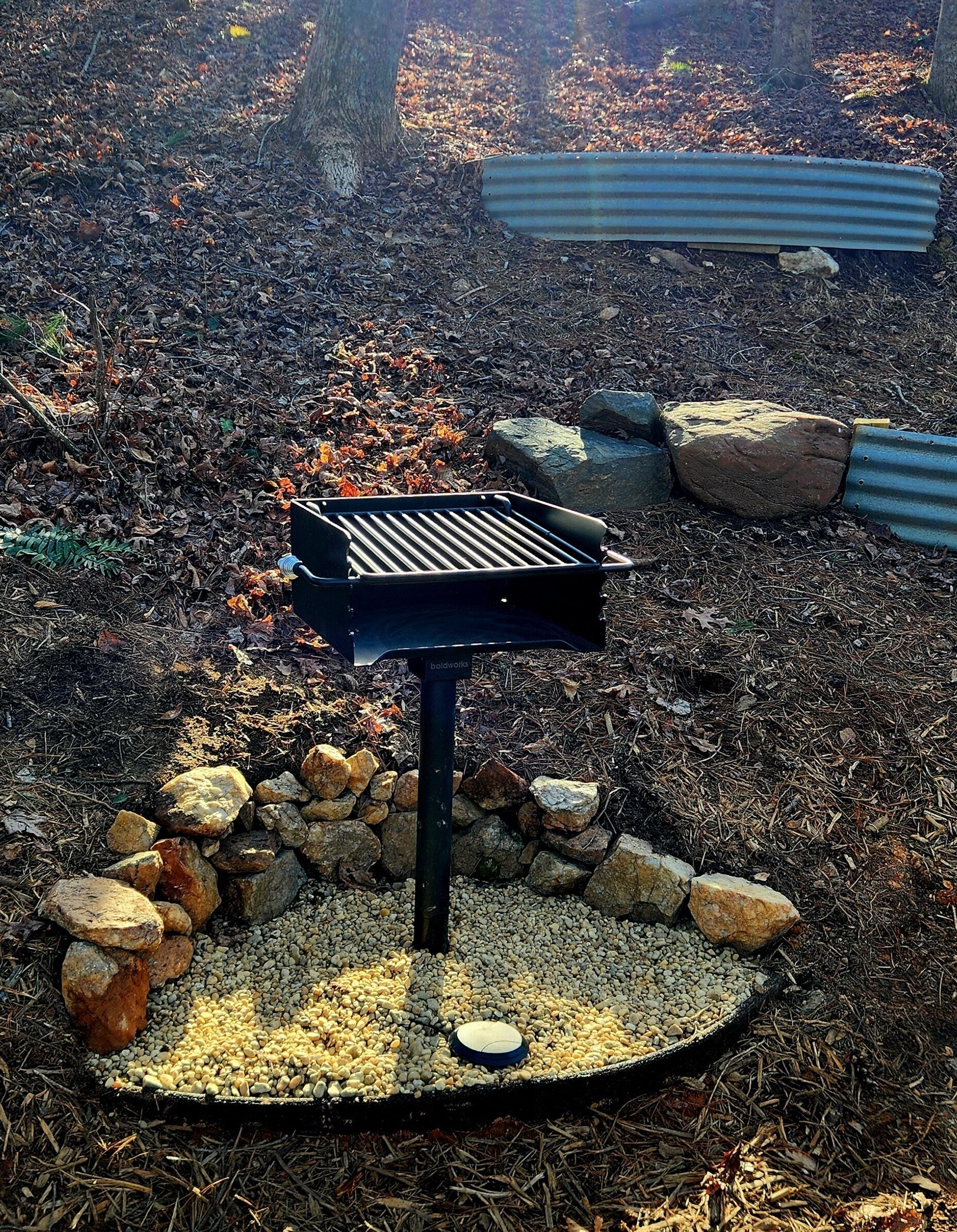 Un barbecue doté d’une grille en métal, entouré d’une zone circulaire recouverte de gravier et de roches, dans un cadre extérieur boisé.