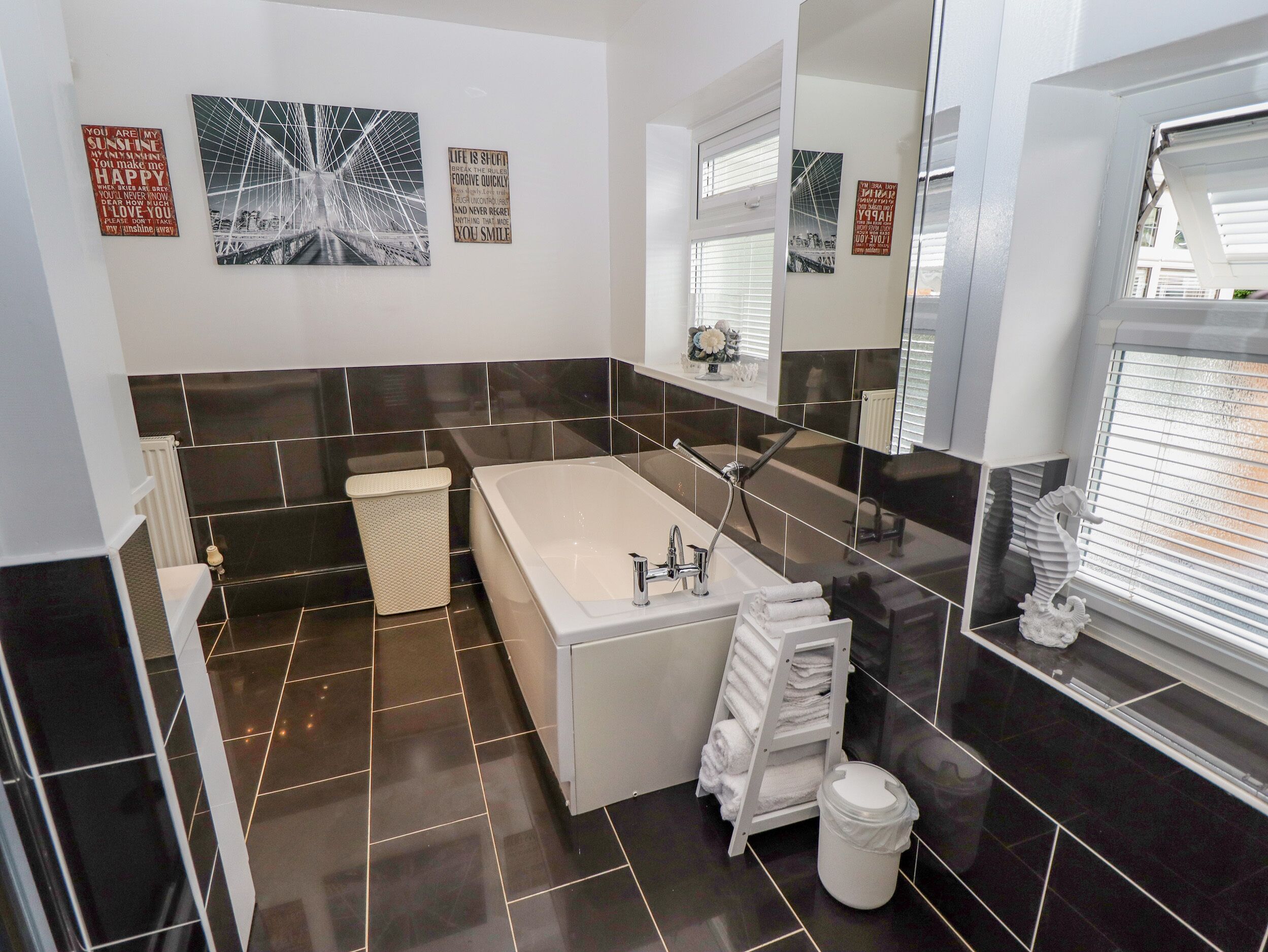 A modern bathroom with a bathtub, a toilet, and a trash bin. There are three wall-mounted pictures and a large mirror.