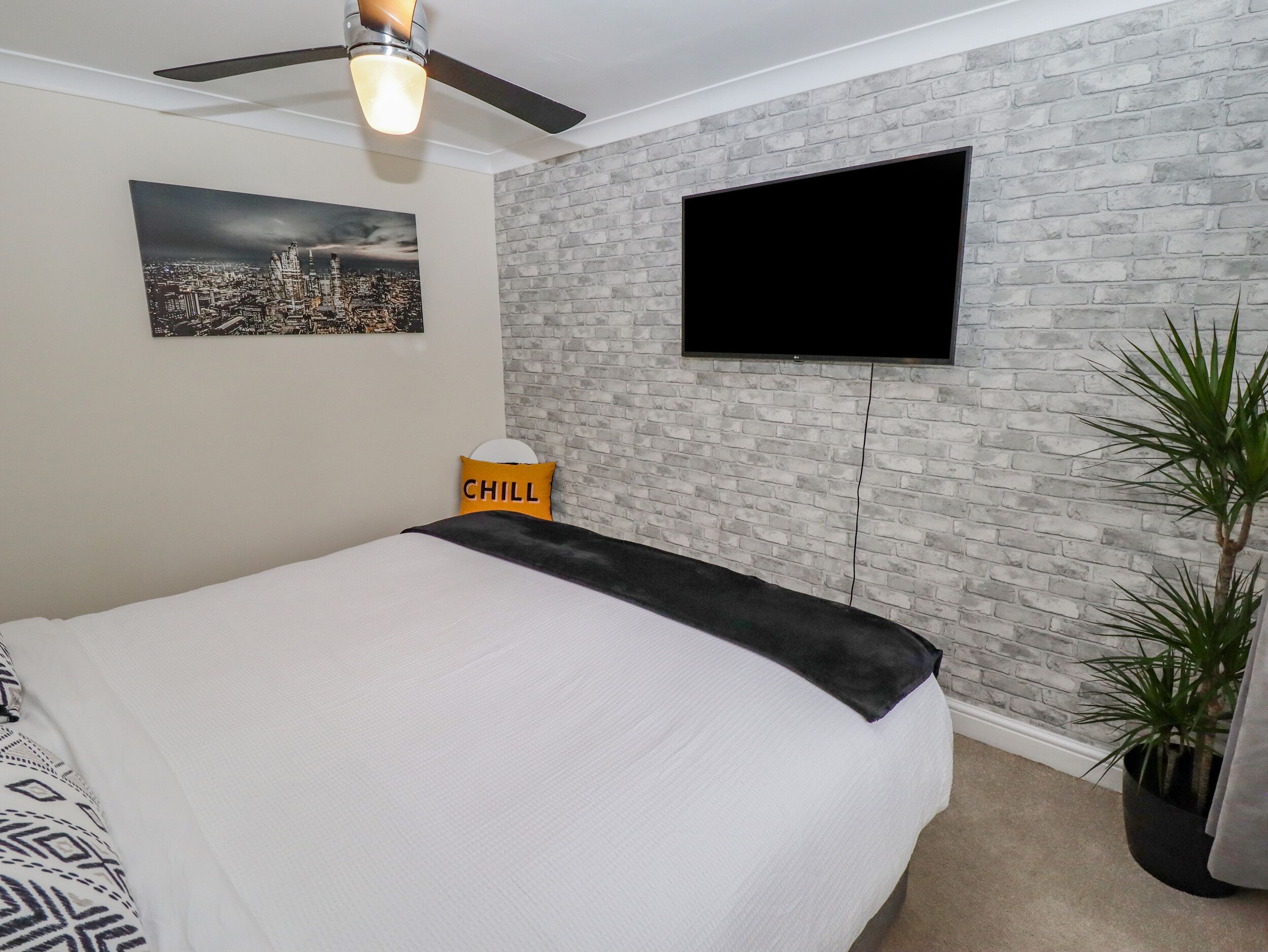 A bedroom with a bed, a cityscape picture, a TV mounted on a brick wall, a ceiling fan, and a plant.