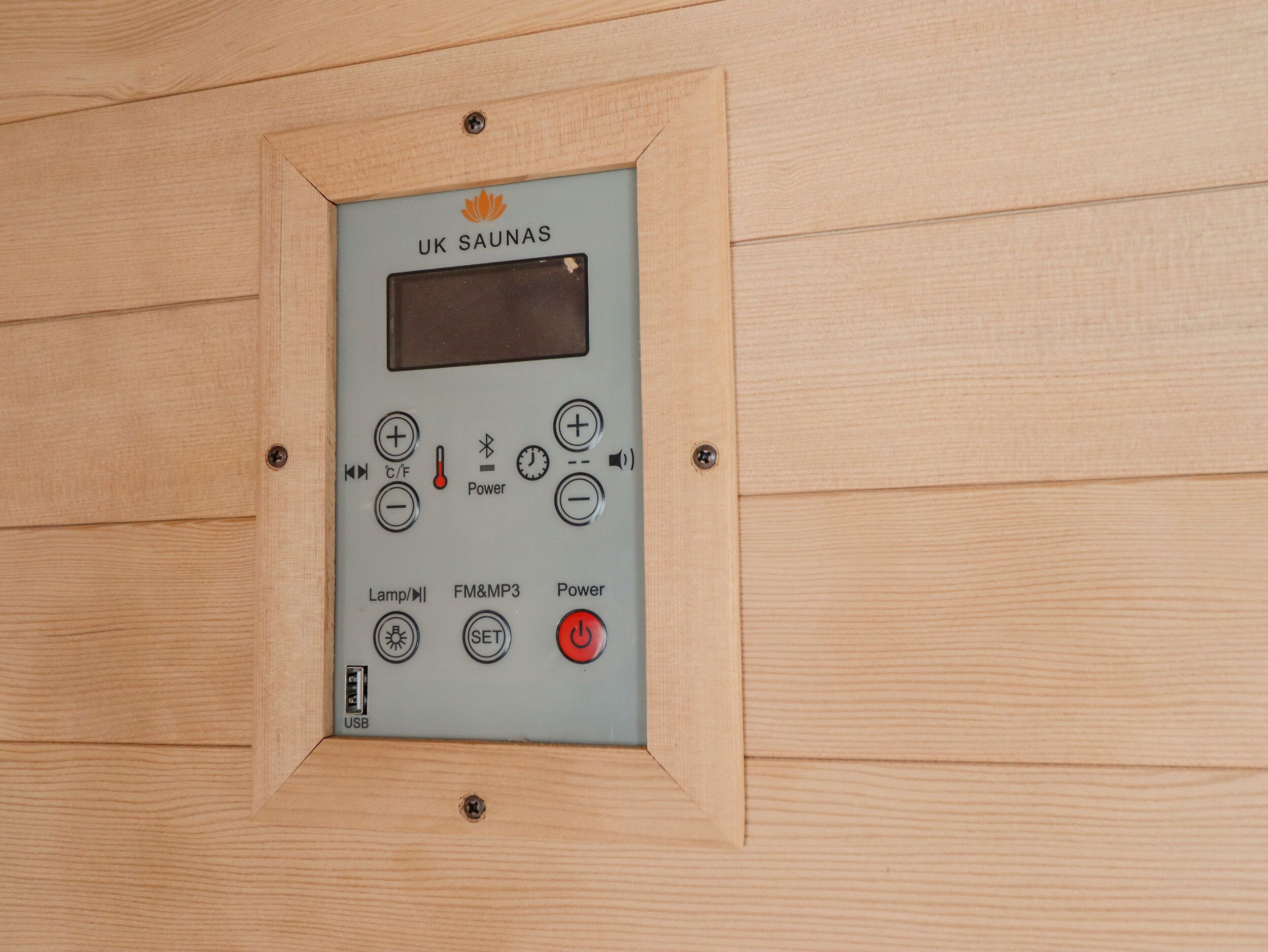 Panel de control de sauna con varios botones y una pantalla digital, fijado a una pared de madera.