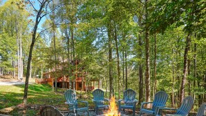 Un foyer entouré de chaises Adirondack, situé dans une zone boisée, avec un chalet en arrière-plan.