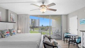 Een hotelkamer met een bed, een plafondventilator, een balkon met uitzicht op de oceaan en een klein zithoekje.