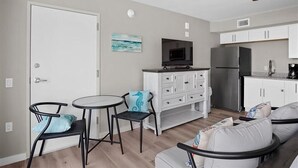 Een ruimtelijk ingerichte leefruimte met een kitchenette, een eethoek met twee stoelen en een tafeltje, een bank en een plafondventilator.
