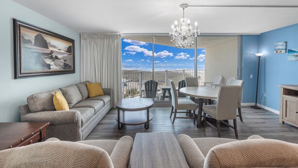 Ein großzügiges Wohnzimmer mit Sofa, Couchtisch und Essbereich. Ein großes Fenster bietet Blick auf den Himmel und einen Balkon.