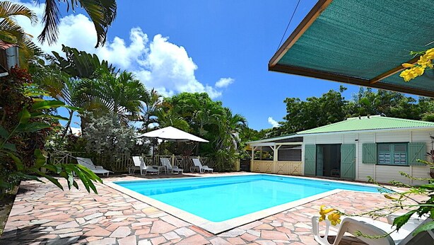 A swimming pool with stone tiles, surrounded by lounge chairs and umbrellas, with a building in the background. - CHARMING HOUSE WITH POOL 5 MINUTES FROM THE BEACH! (LE GOSIER)