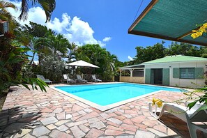 Ein Pool mit Steinfliesen, umgeben von Liegestühlen und Sonnenschirmen, mit einem Gebäude im Hintergrund.
