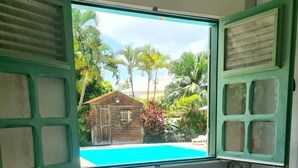 A bedroom with a bed, pillows, a window with a view of a swimming pool and palm trees. - CHARMING HOUSE WITH POOL 5 MINUTES FROM THE BEACH! (LE GOSIER)