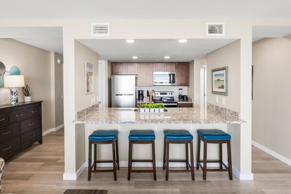 A modern kitchen with granite countertops, stainless steel appliances, and a central island with bar stools. - Sandcastle Condominiums Unit 530 (Port Aransas)
