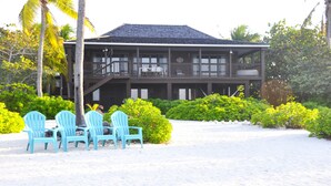 A two-story wooden building with a balcony, surrounded by lush greenery and a sandy beach. - Featherstone - Upstairs Beach Front Apartment (Tibbett's Turn)