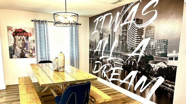 A dining area with a wooden table, benches, and a blue chair. There is a wall with a cityscape mural and a poster with text.