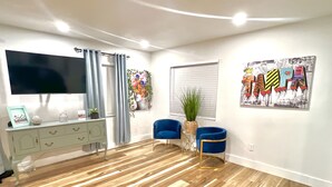 A modern living room with a wooden floor, a flat-screen TV, a white cabinet, two blue armchairs, a small side table, and a colorful artwork on the wall.