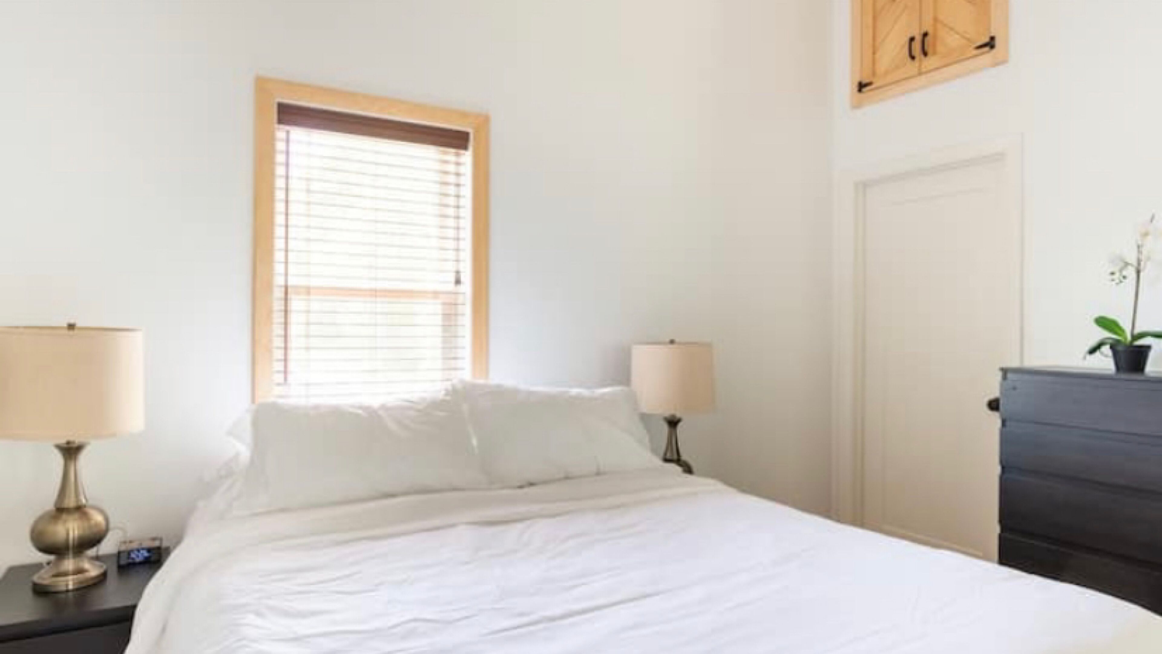 Un dormitorio con cama, dos lámparas de noche, una ventana con persianas, una puerta y un aparador con una planta.