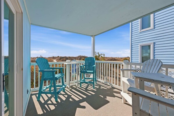 A covered porch with teal and white Adirondack chairs, a small table, and a view of the beach and ocean.