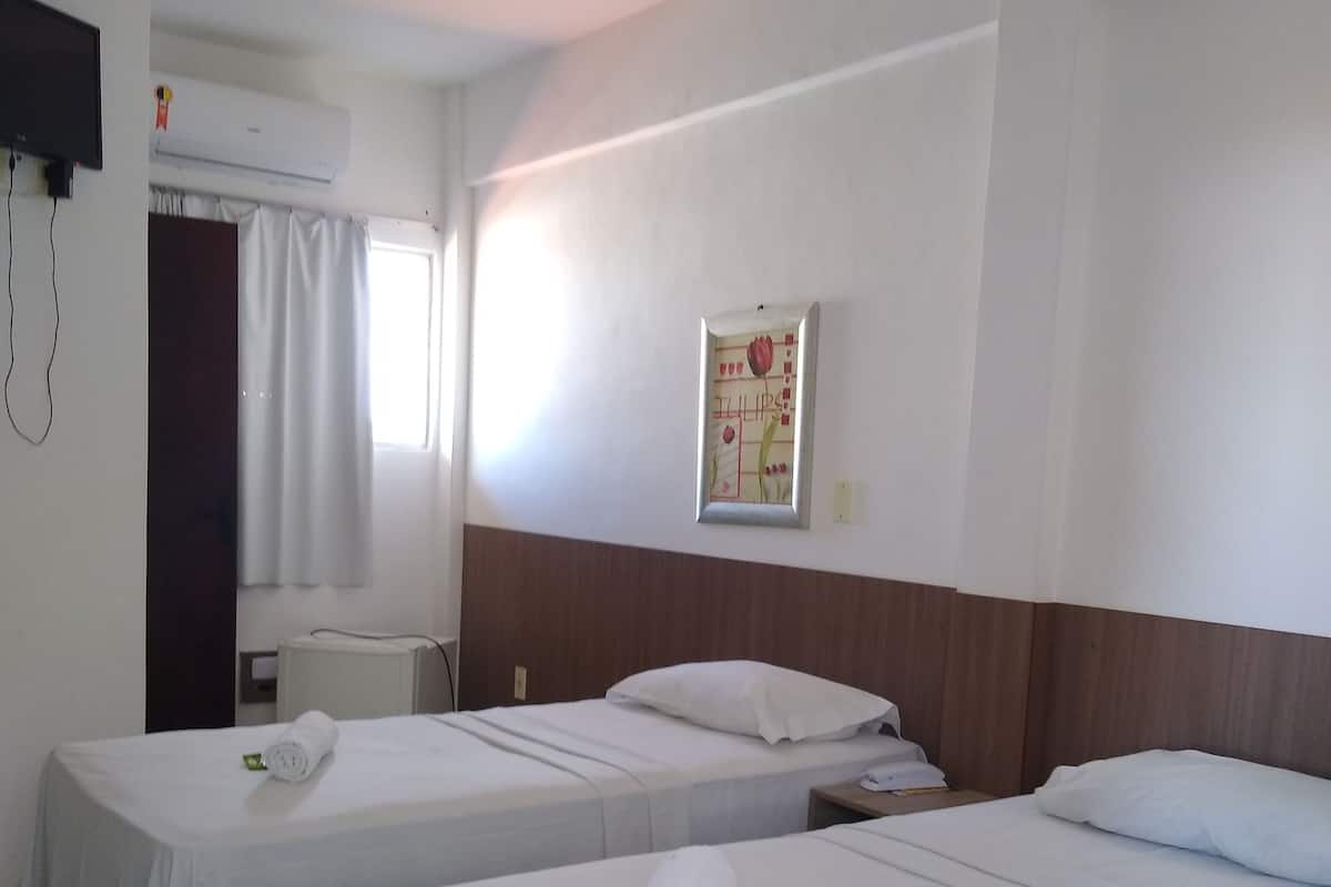 Une chambre d’hôtel avec deux lits simples, une petite table de chevet, un climatiseur fixé au mur et une fenêtre avec des rideaux.