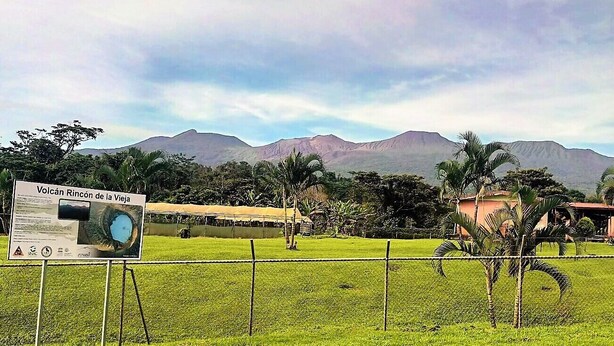 Un cartel de Volcán Rincón de la Vieja con vistas a montañas y un área cercada con césped y palmeras.