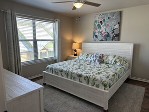 Un dormitorio con cama, mesita de noche, lámpara, ventilador de techo y ventana con cortinas.