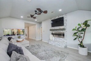 A modern living room with a grey sofa, a large TV, a fireplace, and a ceiling fan.