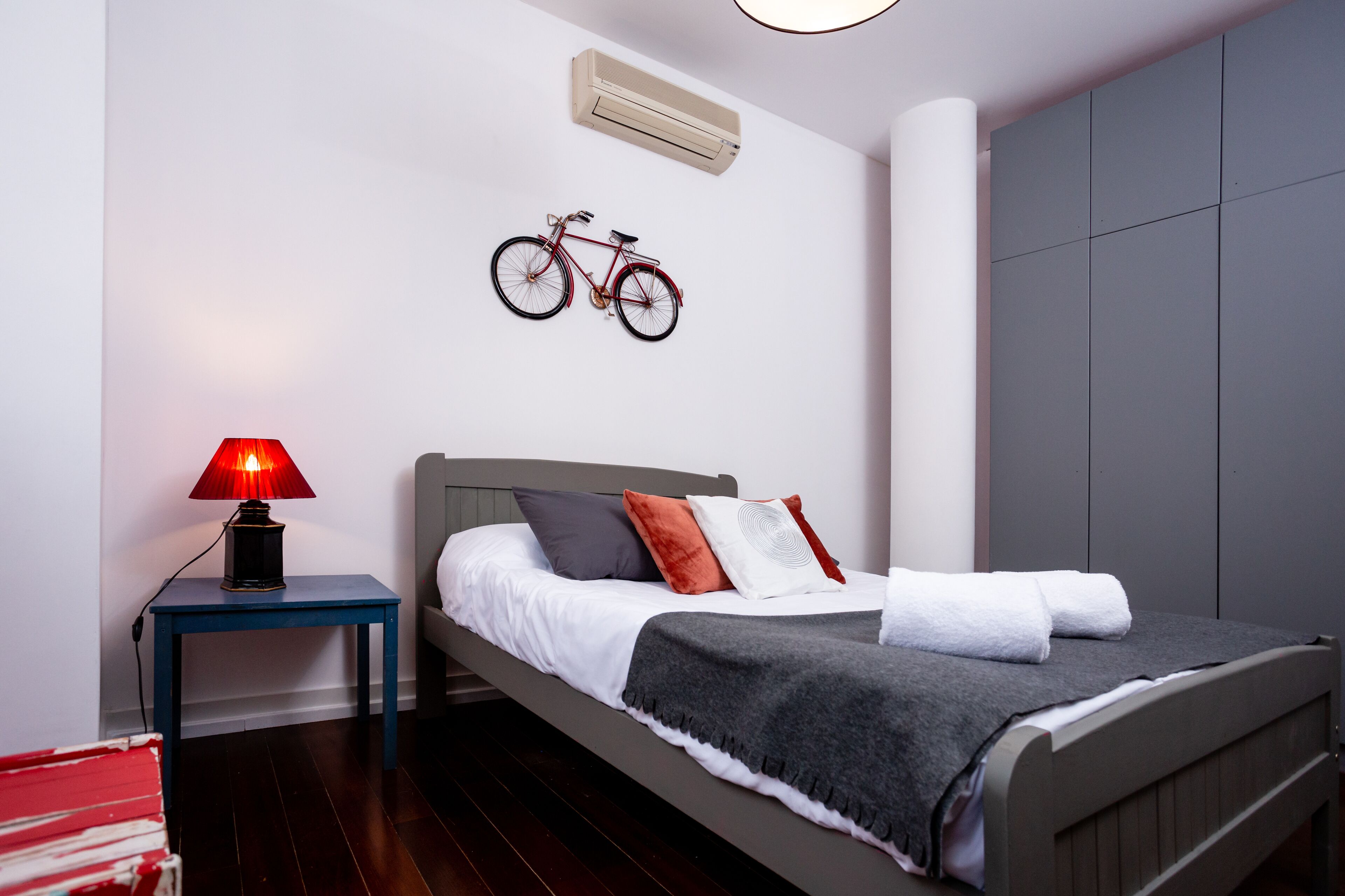 A neatly made bed with a grey headboard, a red bicycle mounted on the wall, and a small table with a red lamp beside it.