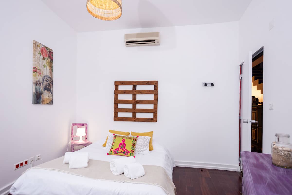 A bedroom with a bed, a nightstand, a wall-mounted air conditioner, a wooden headboard, and a door leading to another room.