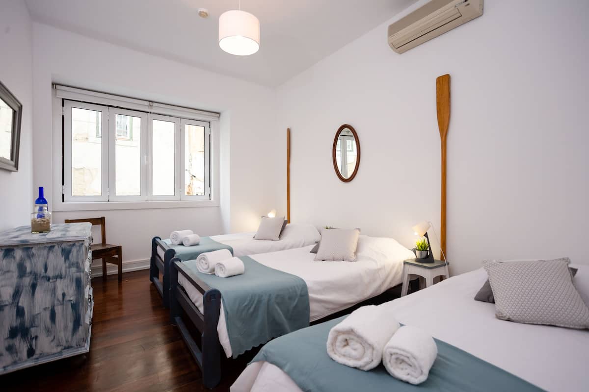 A hotel room with two beds, a small table with a lamp, and a window with blinds.