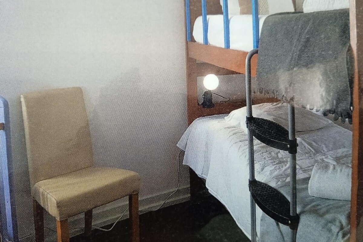 A small, single-bed room with a bunk bed, a chair, and a bedside table with a lamp.