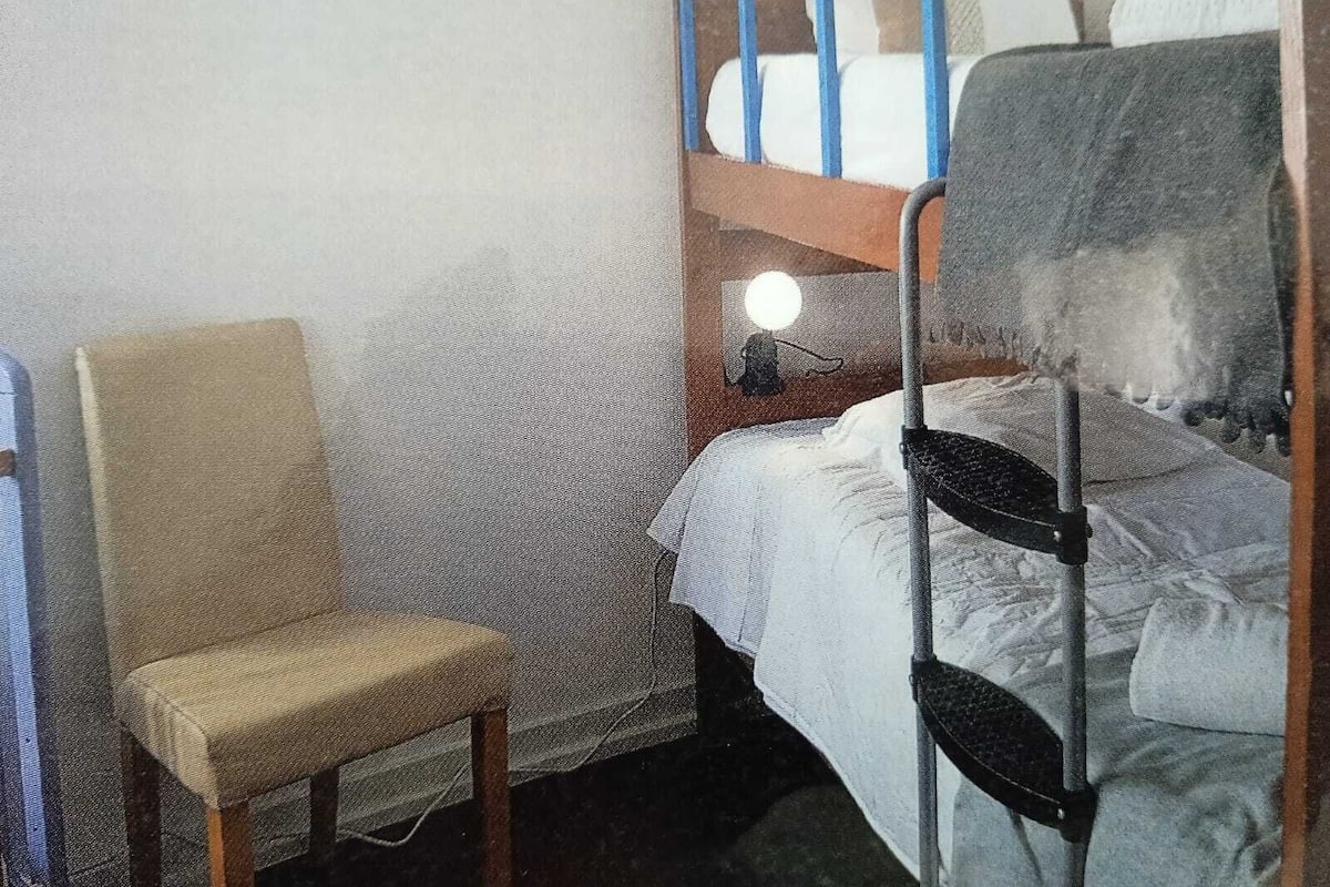 A small, sparsely furnished room with a bunk bed, a single chair, and a bedside table with a lamp.