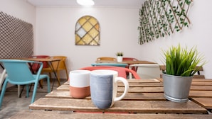 A room with a wooden table, two mugs, a potted plant, and colorful chairs. - Kais Do Sol - Hostel (Seixal)