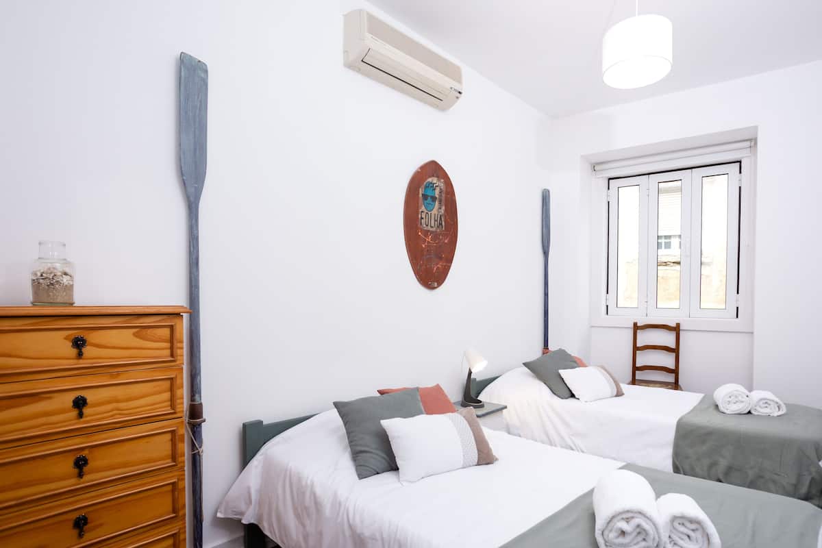 A bedroom with two beds, a wooden dresser, a wall-mounted air conditioner, and a window with a view of the outside.